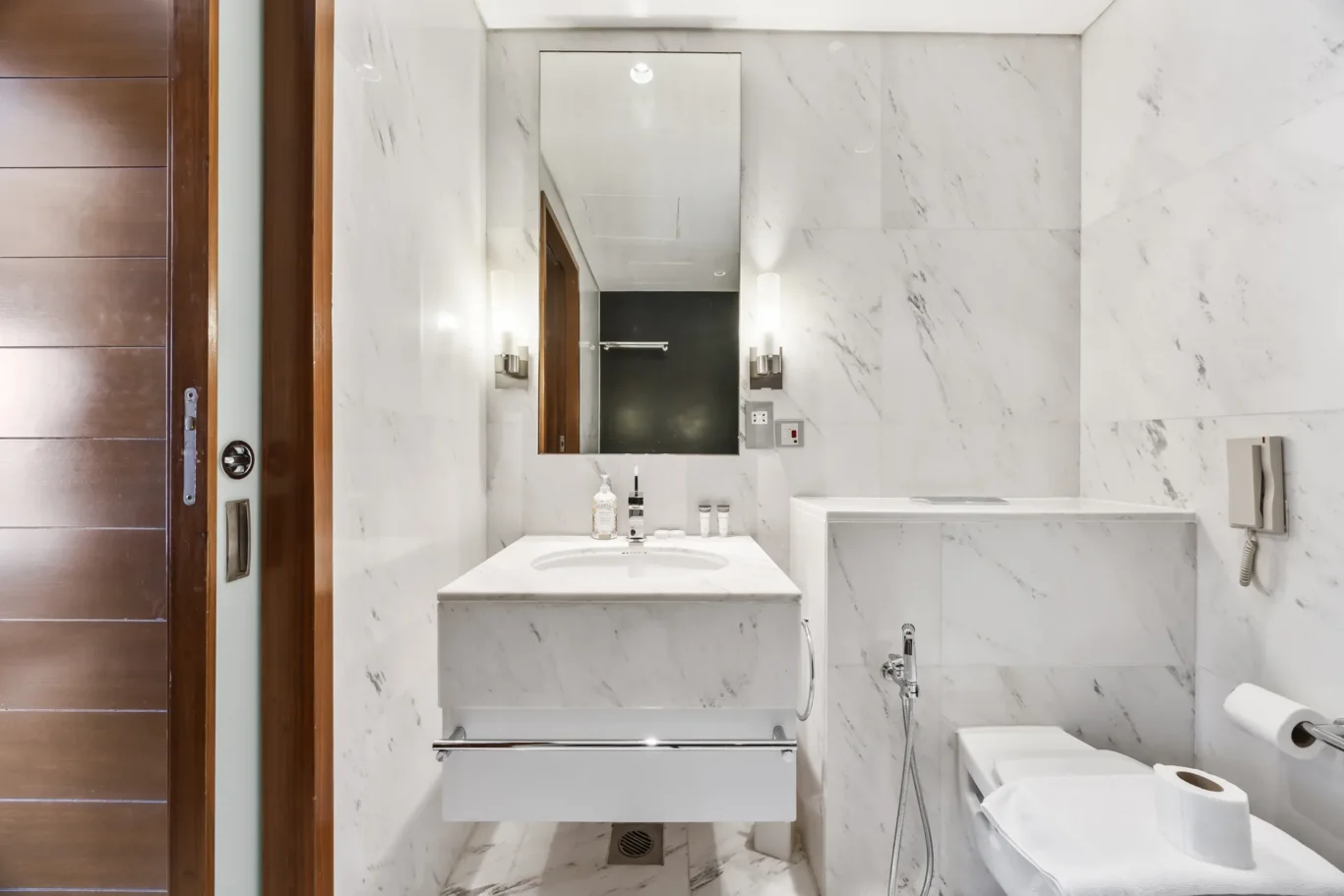 Modern marble bathroom with a wall-mounted sink, large vertical mirror, and toilet on the right; wooden door frame on the left.