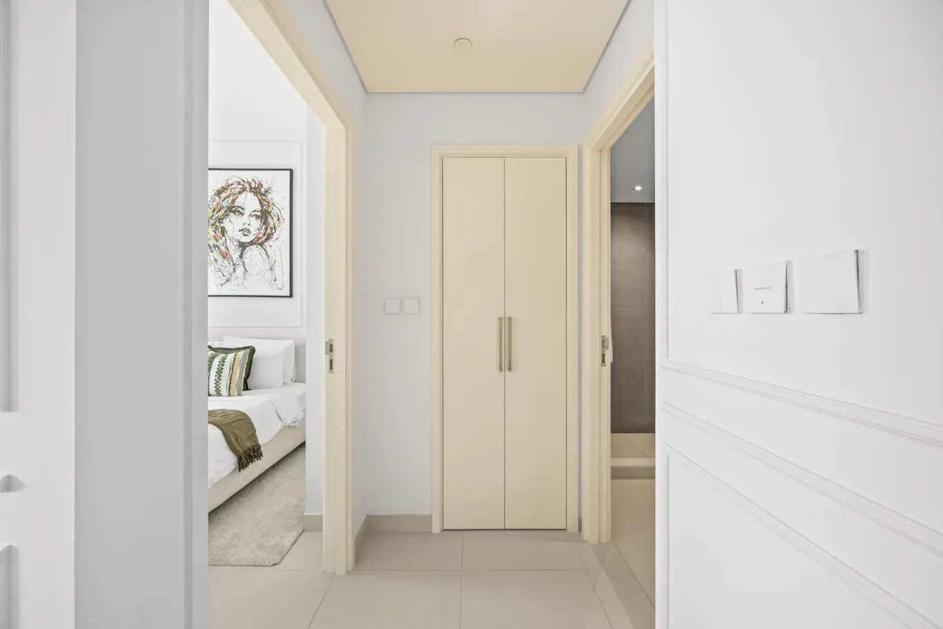 Bright hallway with cream doors; left doorway opens to a bedroom with a framed portrait and pillows, center closet doors, and a right doorway leading to another room.