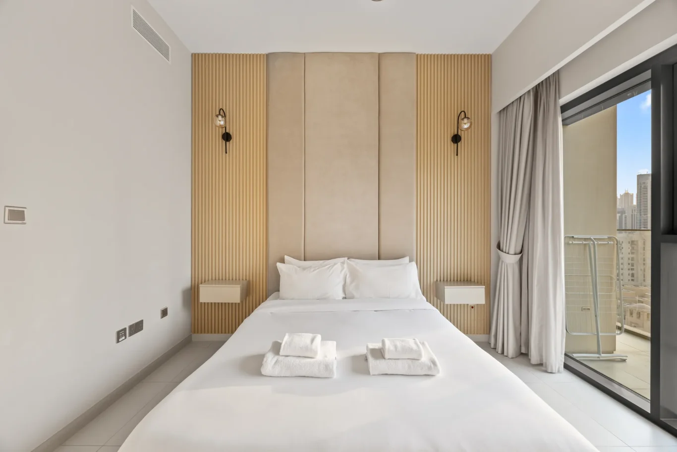 Modern hotel room with a white bed, beige padded headboard, vertical wooden panels, and wall sconces flanking the headboard next to a glass balcony door.