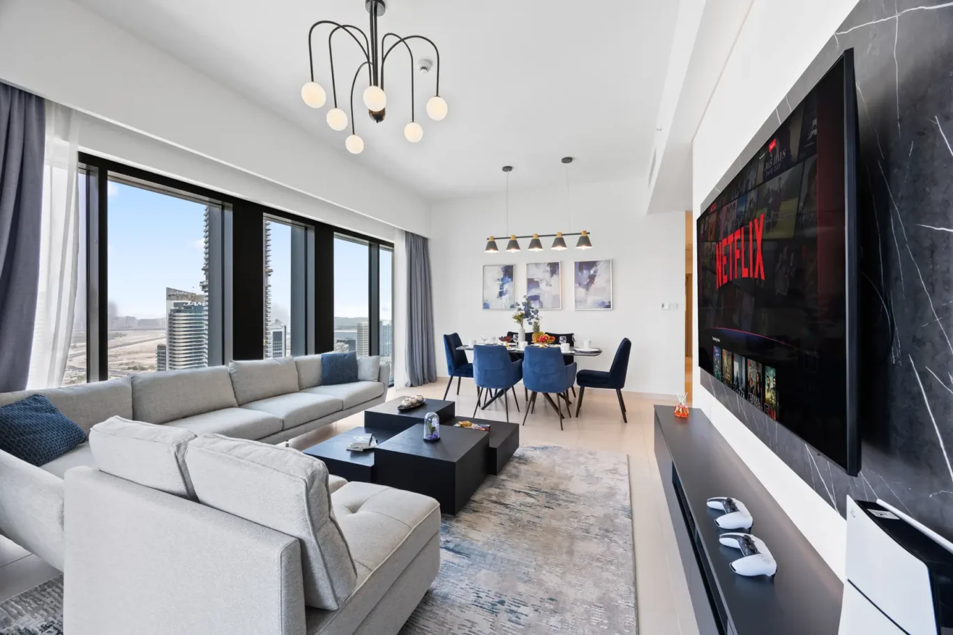 Open-plan living room with a gray sectional sofa, blue dining chairs, and a large wall-mounted Netflix screen next to a marble accent wall, with city views outside the windows on the left.