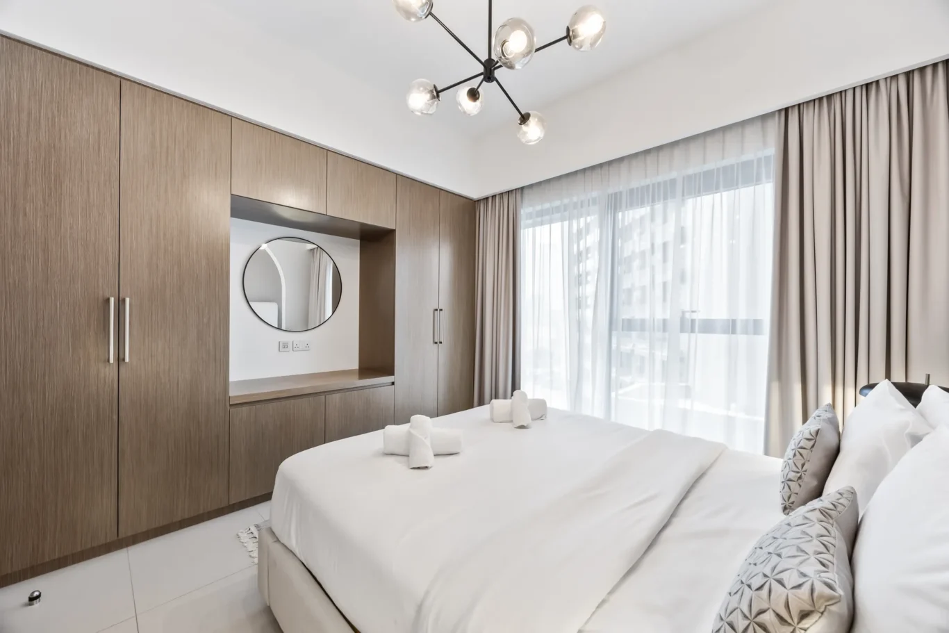 Modern hotel bedroom with wood-toned built-in wardrobes, a round wall mirror, and a geometric ceiling chandelier over white bedding.",