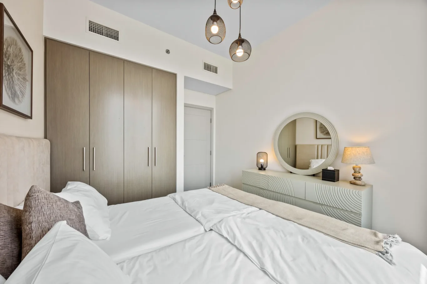 Modern bedroom with a large wooden wardrobe, round mirror above a patterned dresser, and bedside lamps on a light wall.”]} {