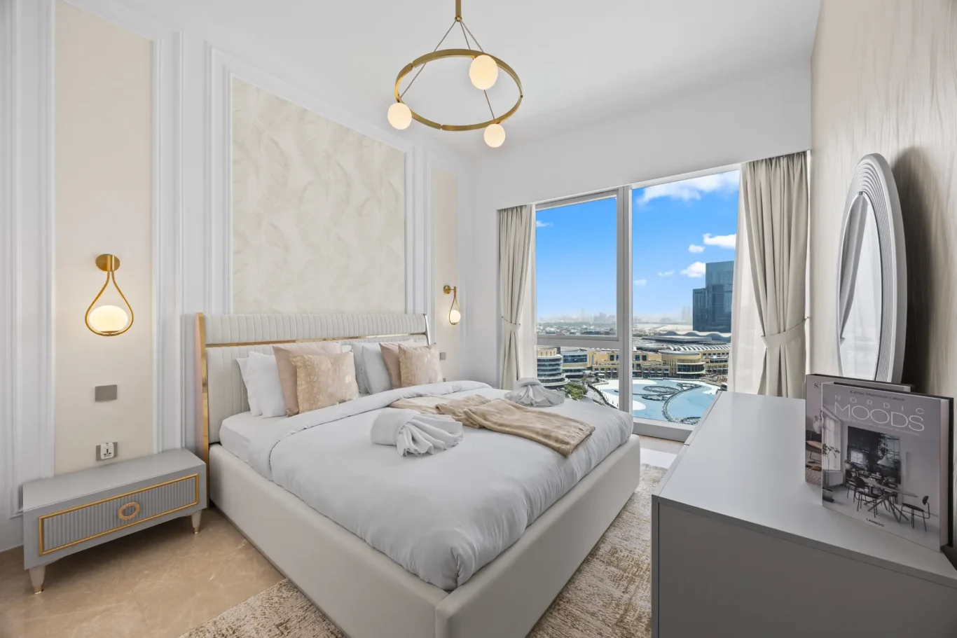 Luxurious hotel bedroom with a plush bed, pastel bedding, and gold-accent nightstands facing a large window overlooking a city skyline.