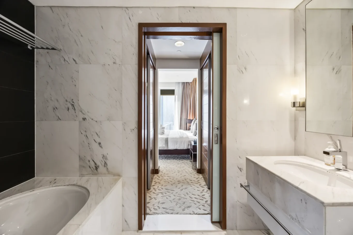 View from a marble bathroom into a bedroom through a wooden-framed doorway, bed with white linens visible beyond