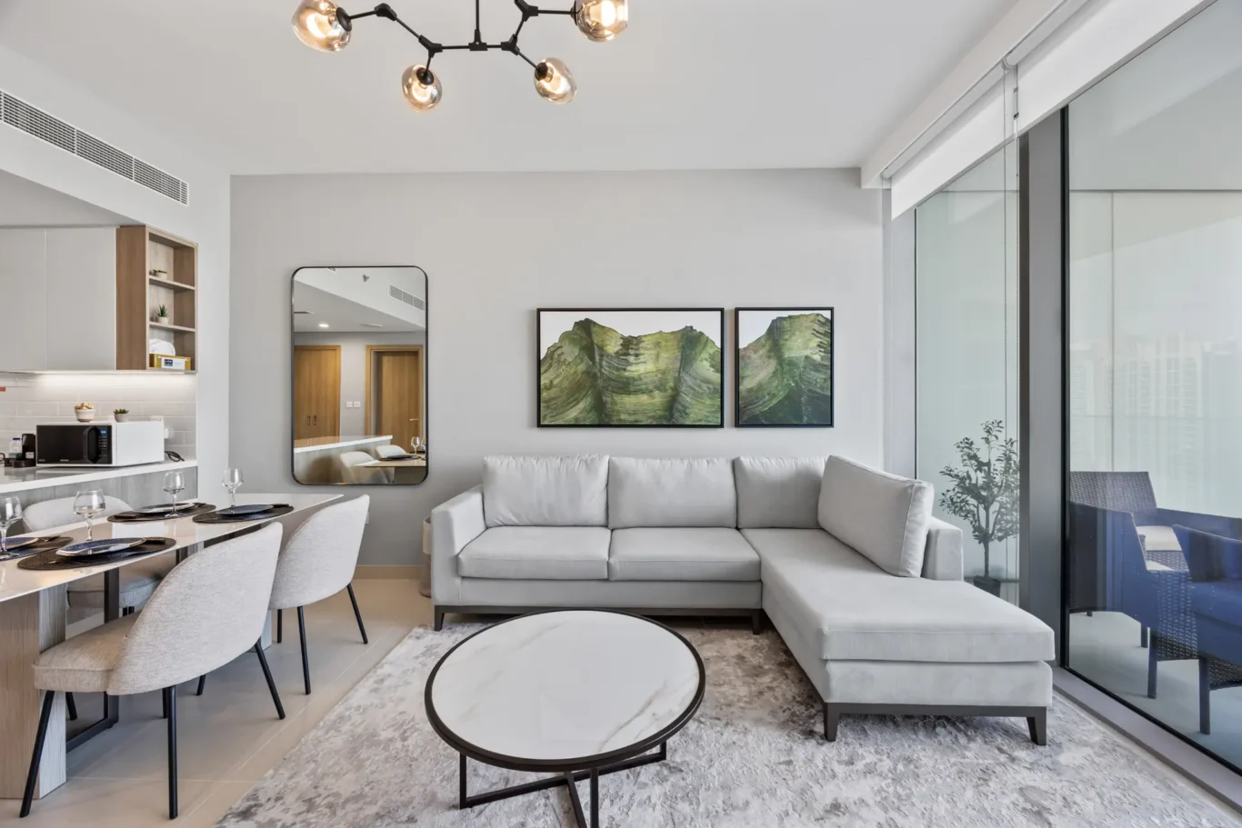 Bright modern living area with a gray L-shaped sofa, two framed green landscape prints, and a glass balcony door to the outside terrace.