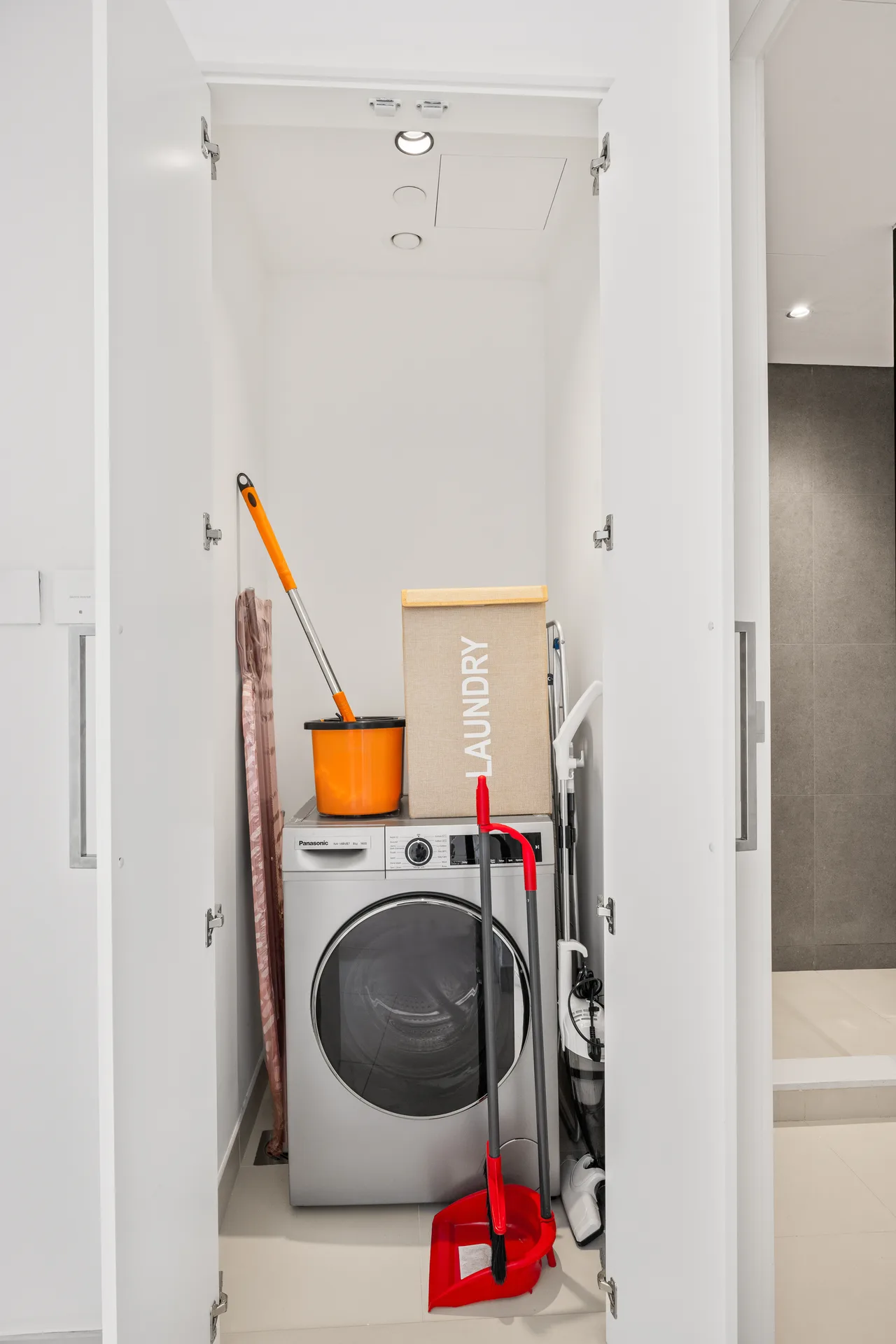 Open laundry closet containing a front-loading washer and dryer, a beige 'LAUNDRY' box, an orange mop bucket, and cleaning tools/levers.