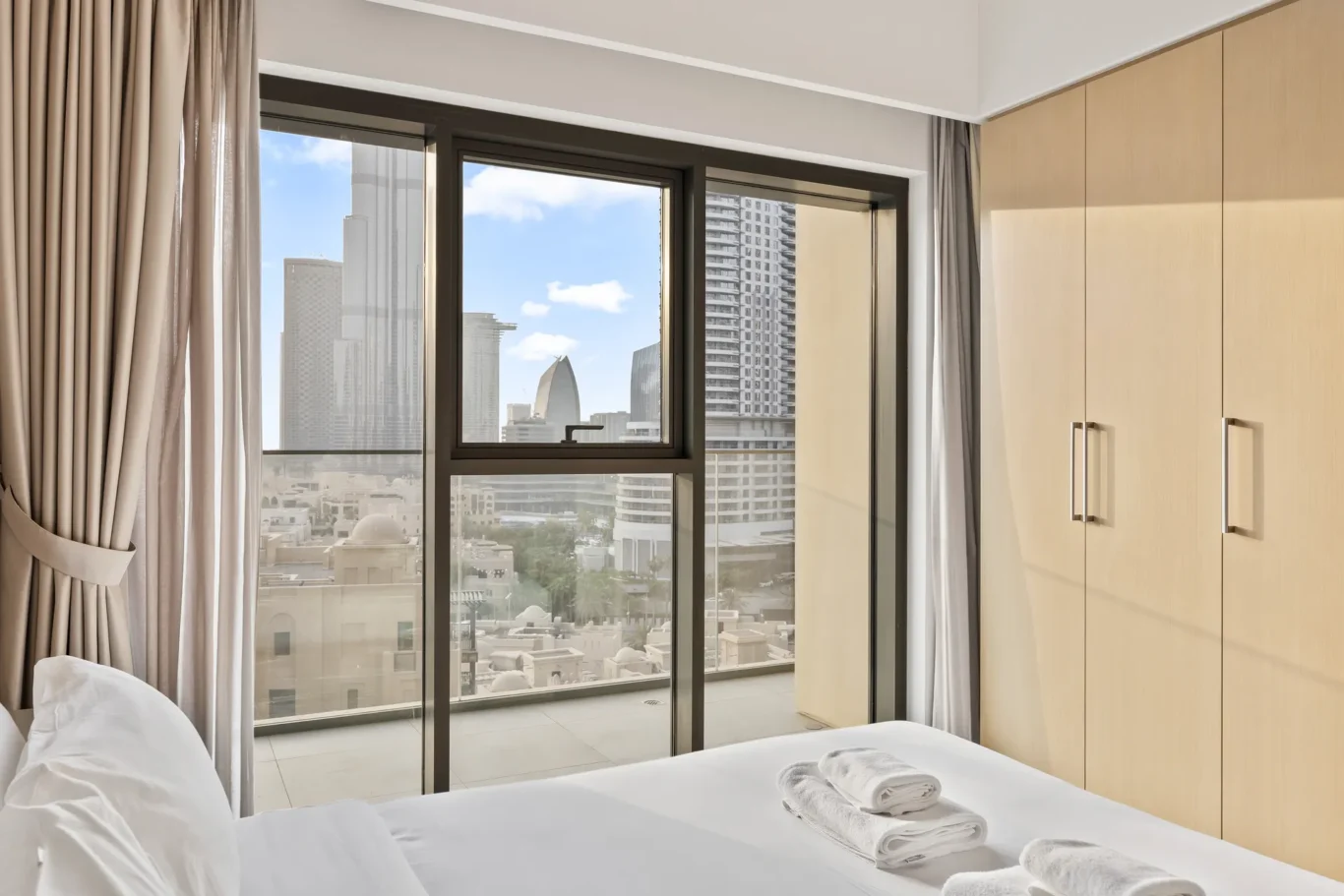 Hotel bedroom with a large window overlooking the city skyline, a made bed in the foreground, beige curtains, and light wooden wardrobes along the right.