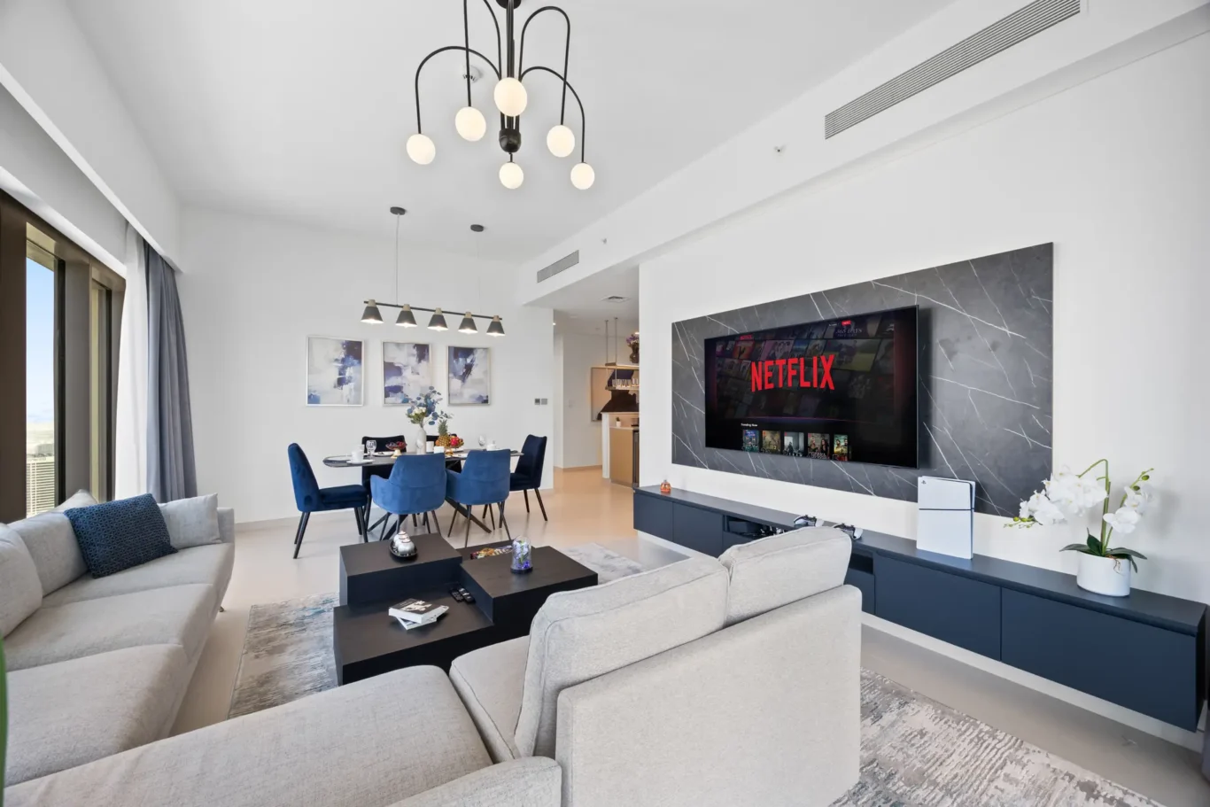 Bright, modern open-plan living and dining area with gray sofas, blue dining chairs, and a wall-mounted Netflix TV on a marble panel.
