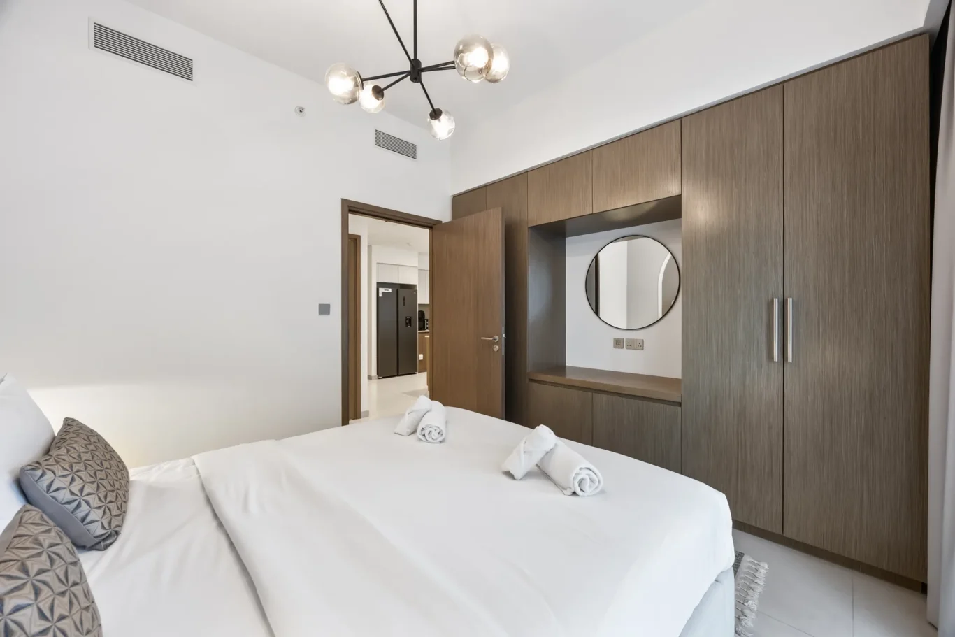 Modern hotel bedroom with white bed linens, decorative gray pillows, and two rolled towels on the foot of the bed; large wooden built-in wardrobe with a circular mirror.