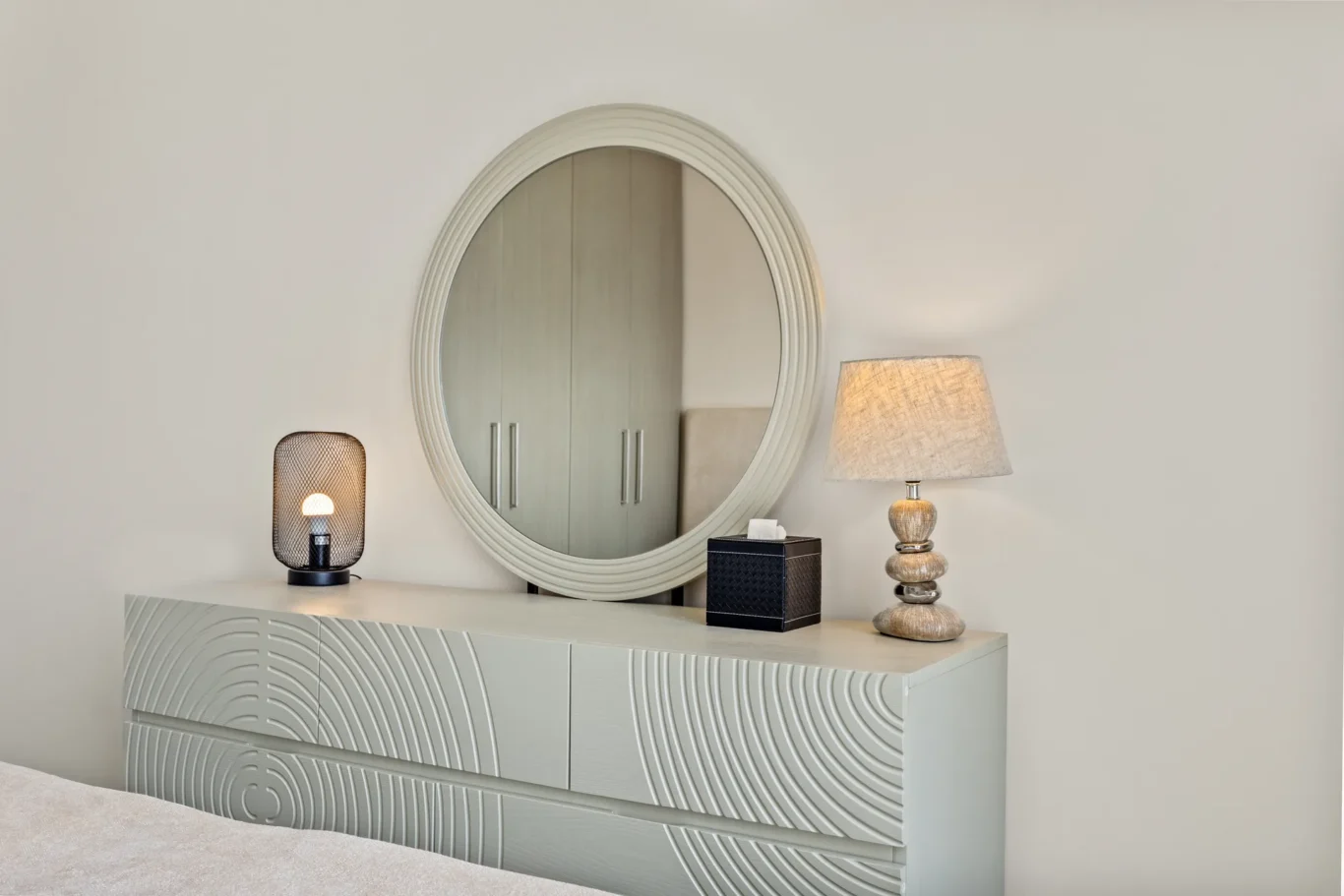 Round, light-gray dresser with a large circular mirror above it, two lamps, and decorative storage boxes in a neutral-toned bedroom setup