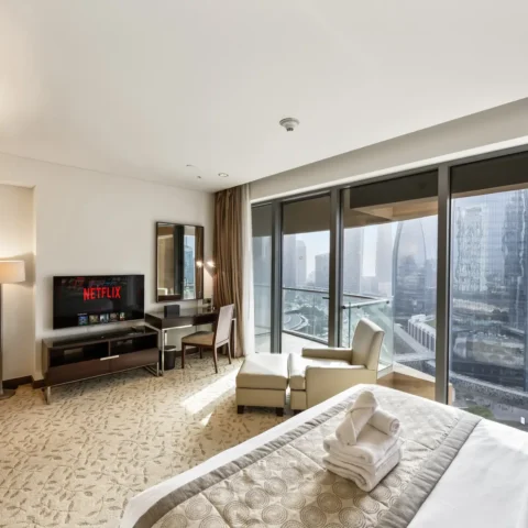 Hotel room with a king bed, seating area, and floor-to-ceiling windows showing a city skyline outside, plus a Netflix TV on a low console.
