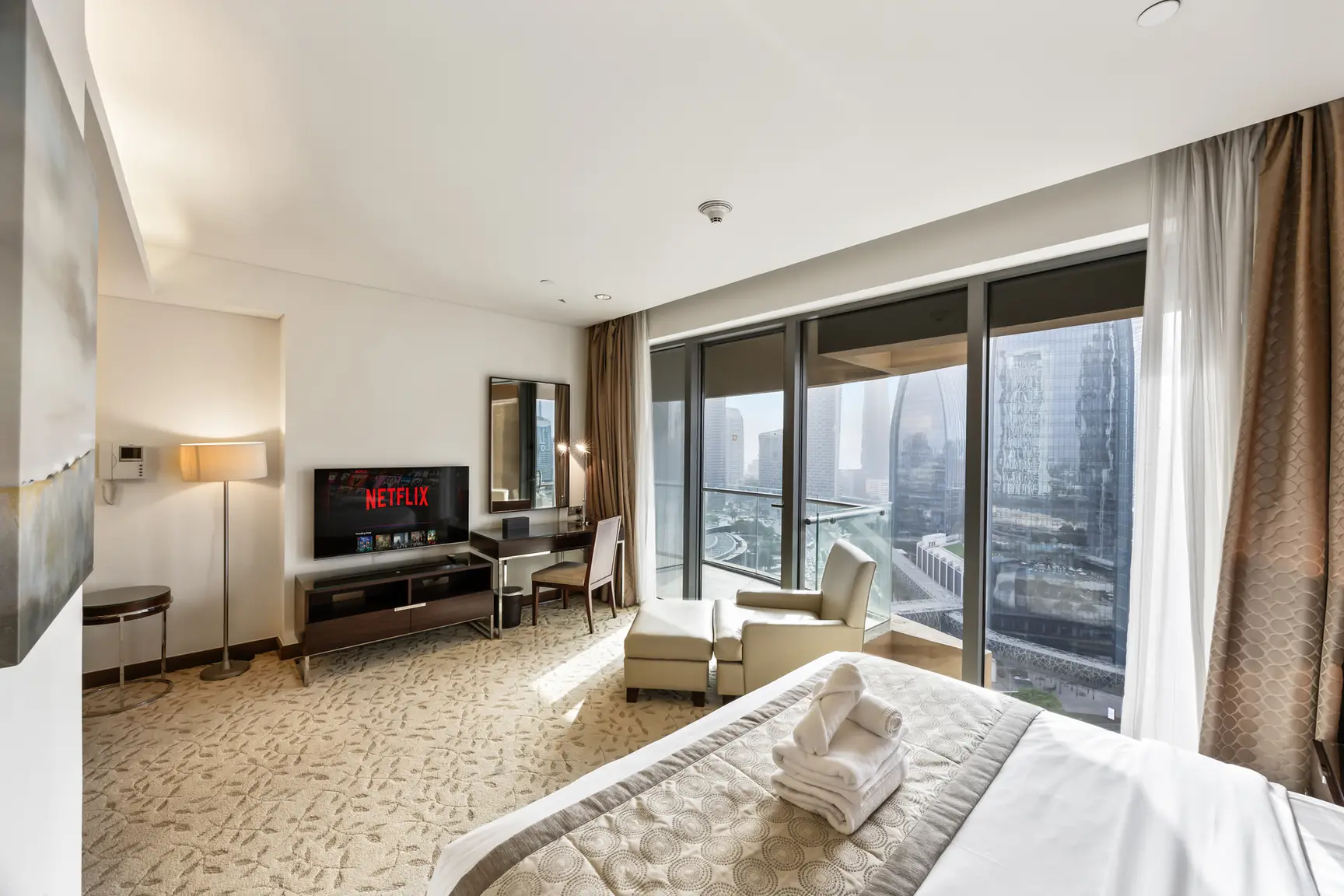 Hotel room with a king bed, seating area, and floor-to-ceiling windows showing a city skyline outside, plus a Netflix TV on a low console.