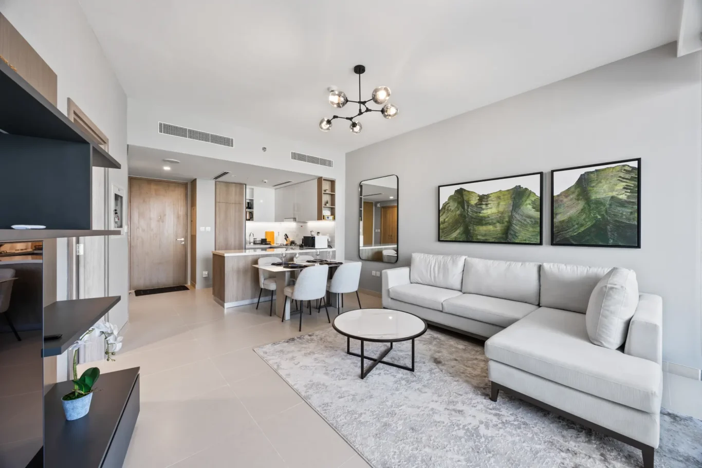Open-plan living area with a light gray sectional sofa, two framed green mountain artworks, and a round coffee table.