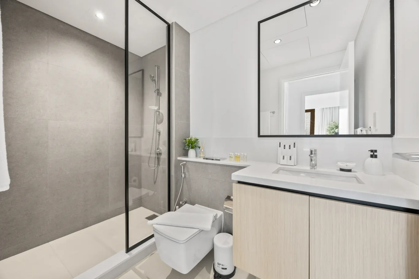 Modern bathroom with glass-enclosed shower, gray tiled walls, and a white countertop sink with a large framed mirror above the vanity.