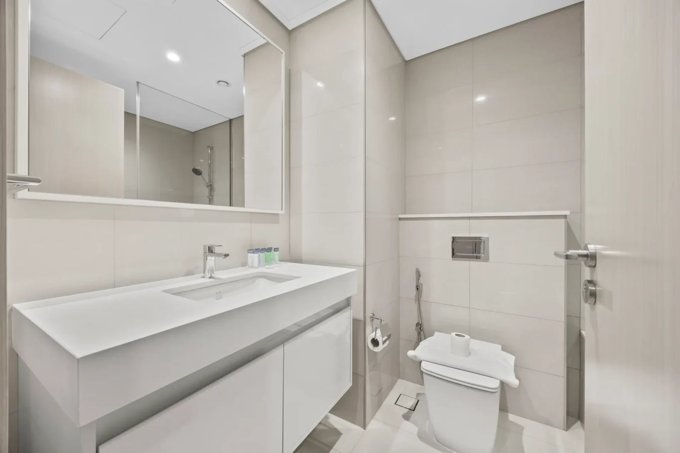 Modern white bathroom with a rectangular sink, large wall mirror, and chrome fixtures; toilet and shower visible in tiled space.