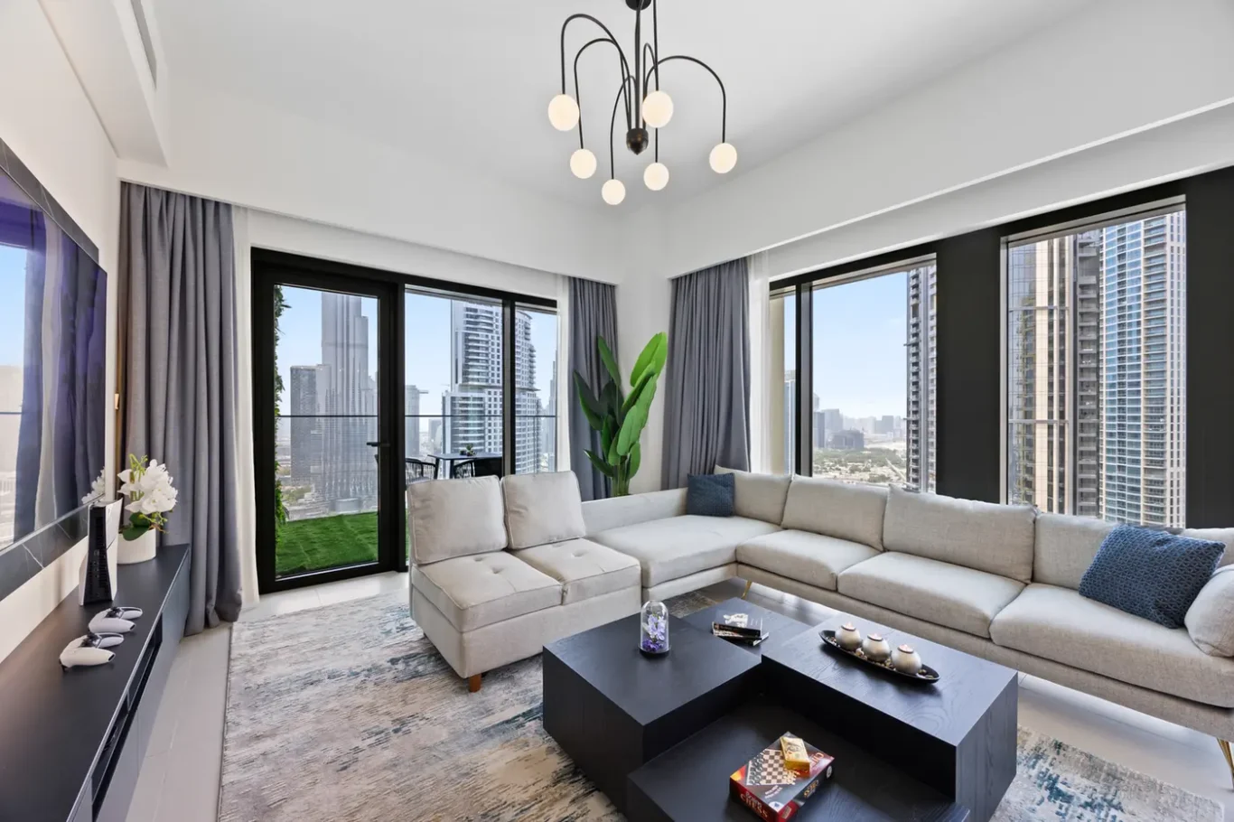 Modern living room with a grey sectional sofa, black coffee tables, and large floor-to-ceiling windows looking out at a city skyline.