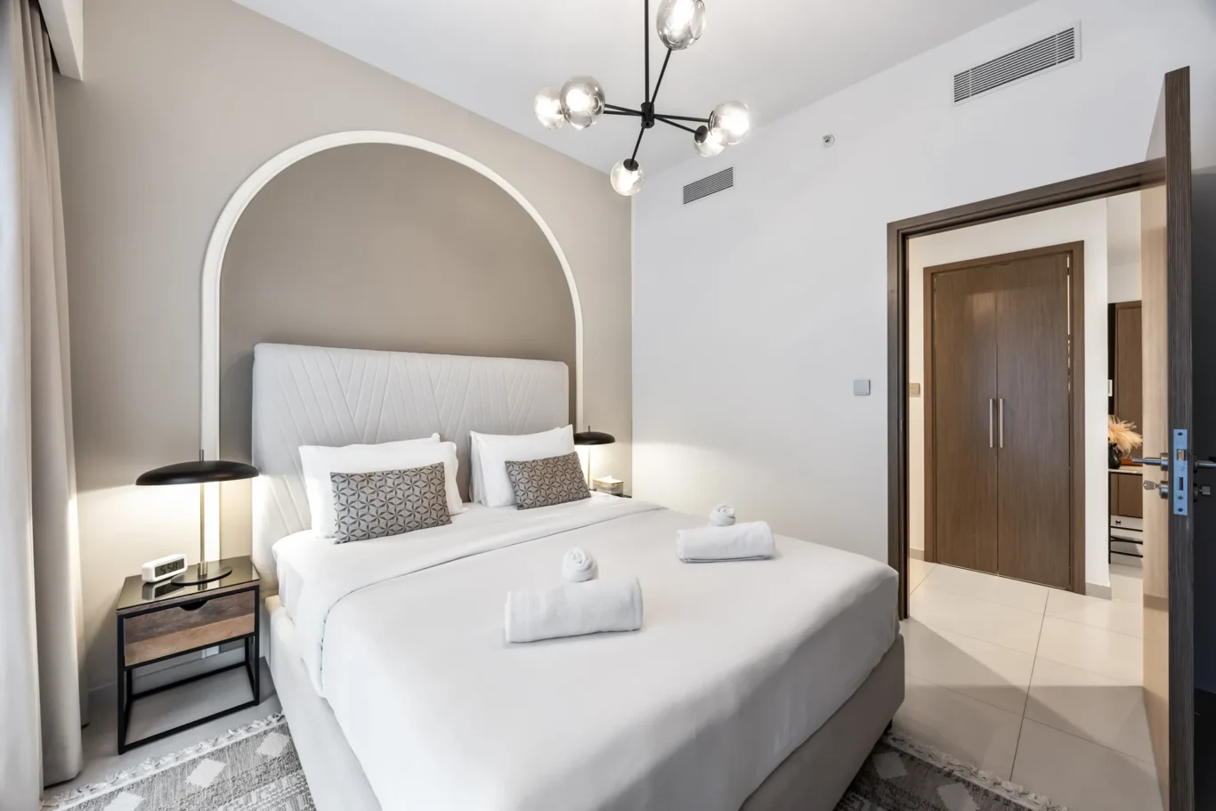 Modern hotel bedroom with a large bed, white linens, and a curved, light-gray headboard set against a taupe accent wall.