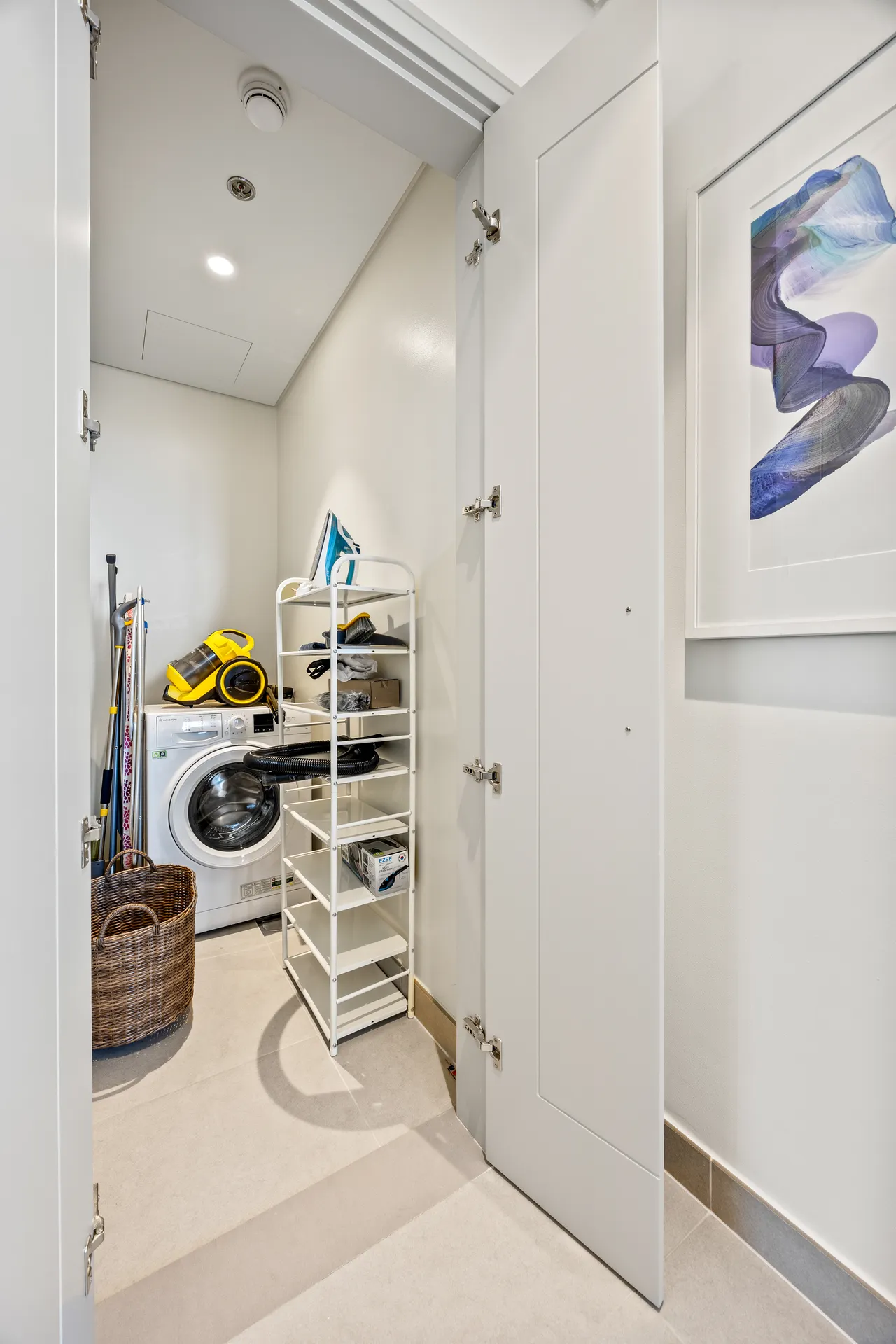 Small laundry closet with a front-loading washing machine, white shelves, and cleaning supplies organized on a rack and floor baskets nearby.