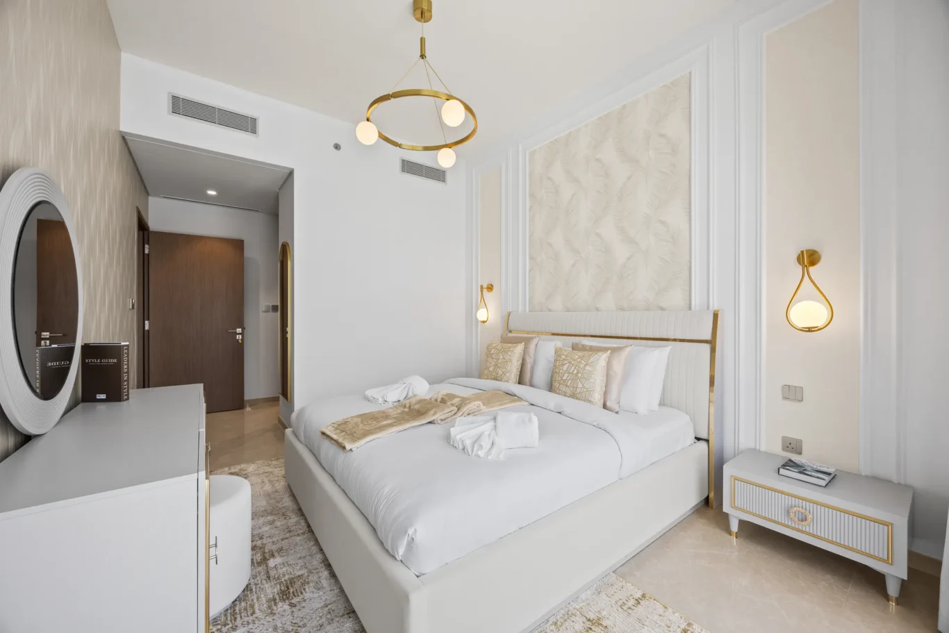 Luxurious hotel bedroom with white and gold decor, large bed, circular gold chandelier, mirrored dresser, and bedside lamps with plush pillows.