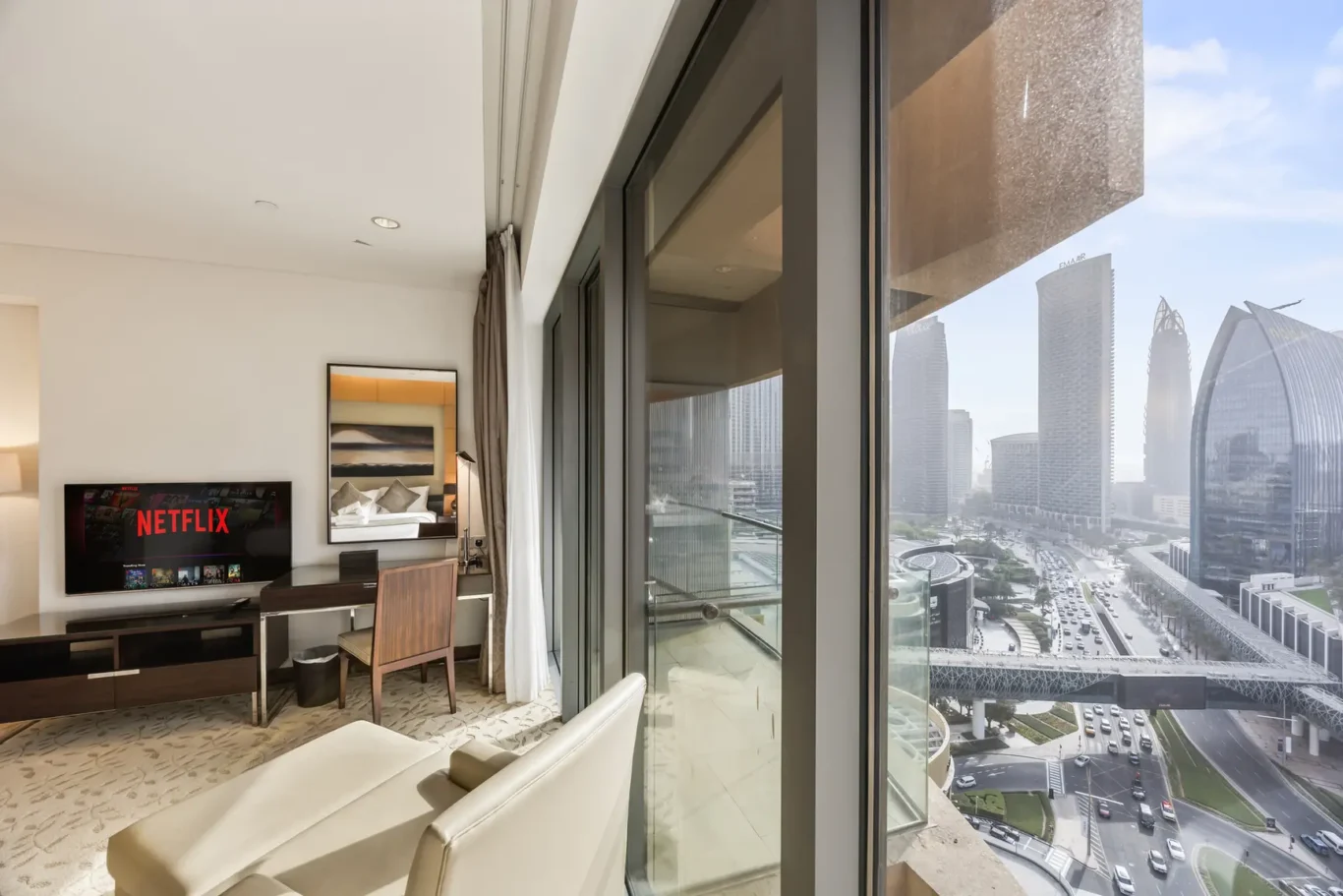 Hotel living area with a wall-mounted Netflix TV, sofa, and desk facing large glass doors to a city view.