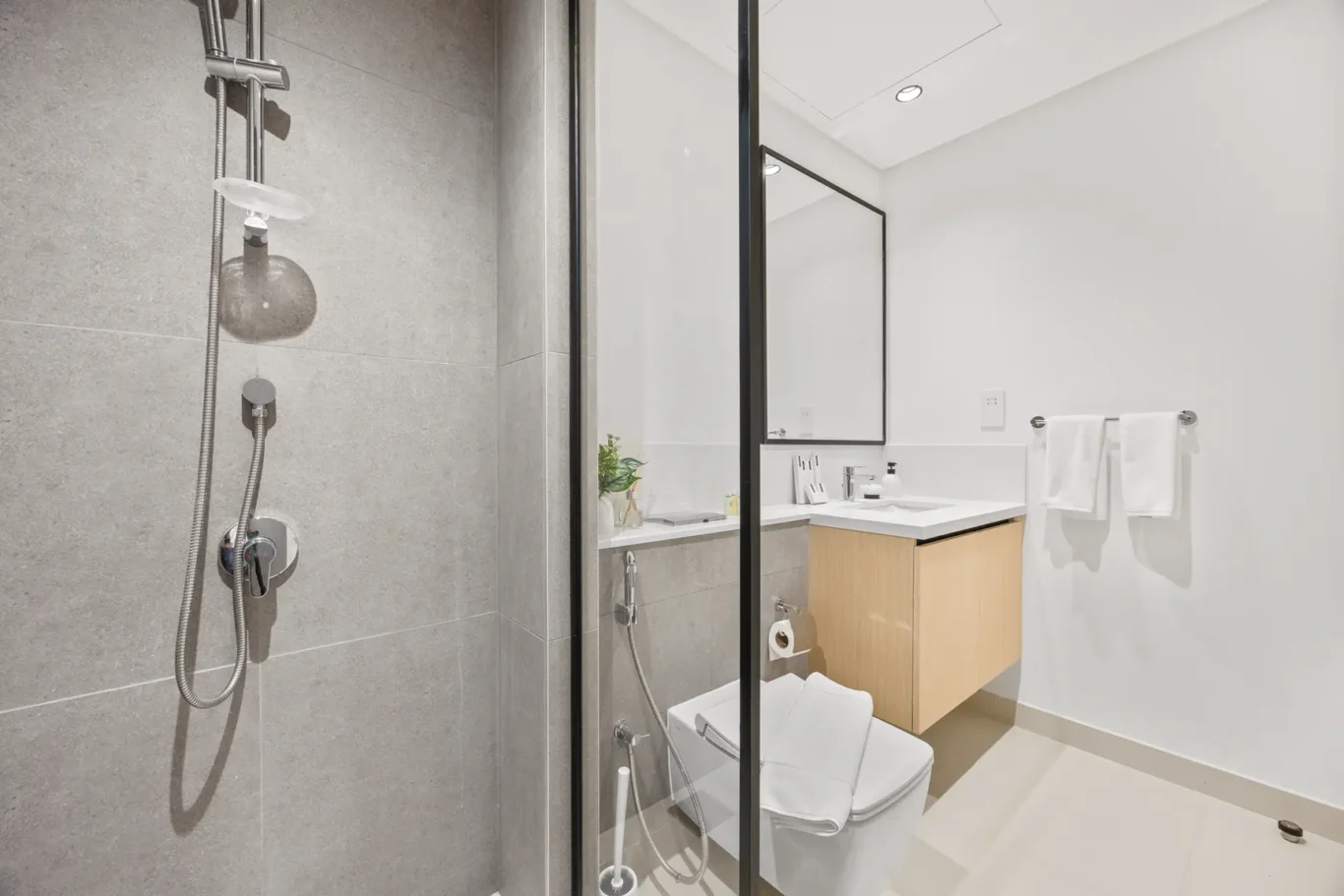 Modern bathroom showing a gray tiled shower area with a glass partition, white sink with wooden vanity, large rectangular mirror, and white towels on a rack