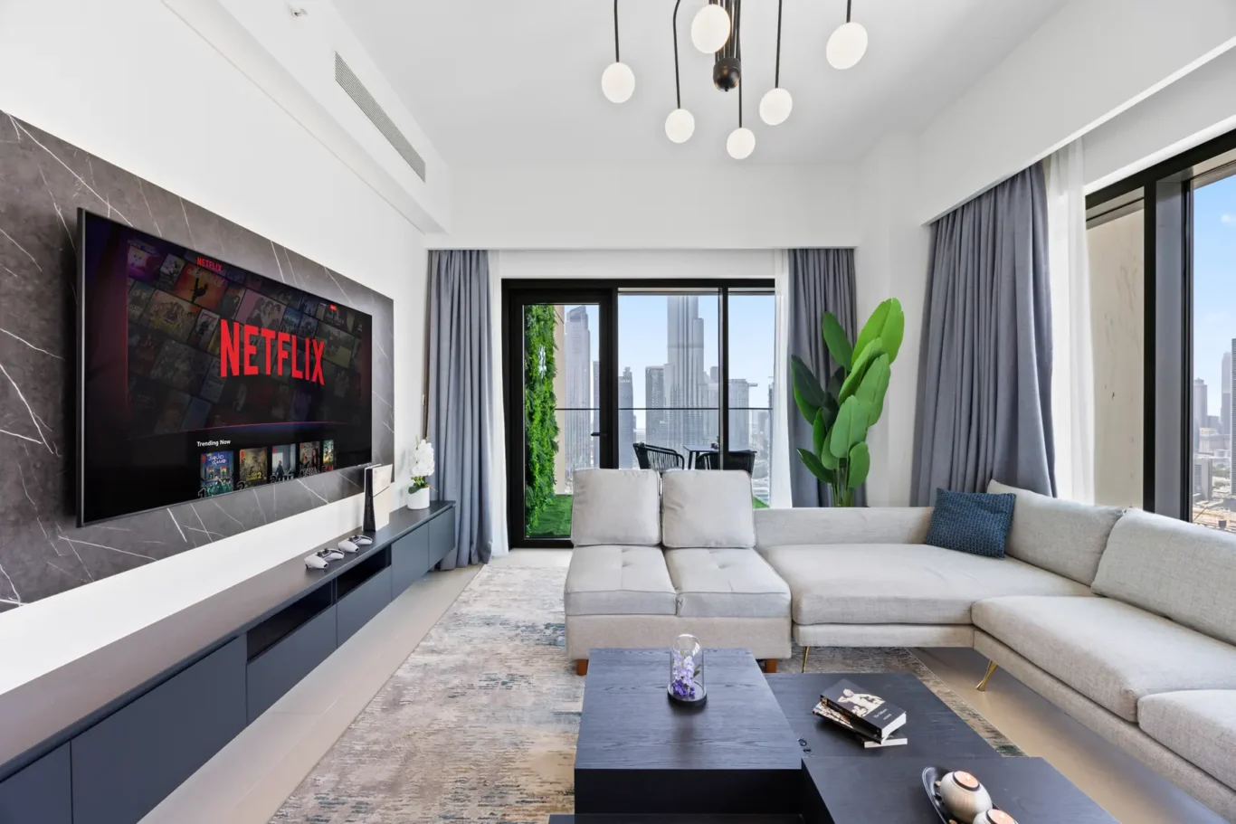 Modern living room with a wall-mounted Netflix TV, gray sectional sofa, and glass balcony doors overlooking a city skyline.