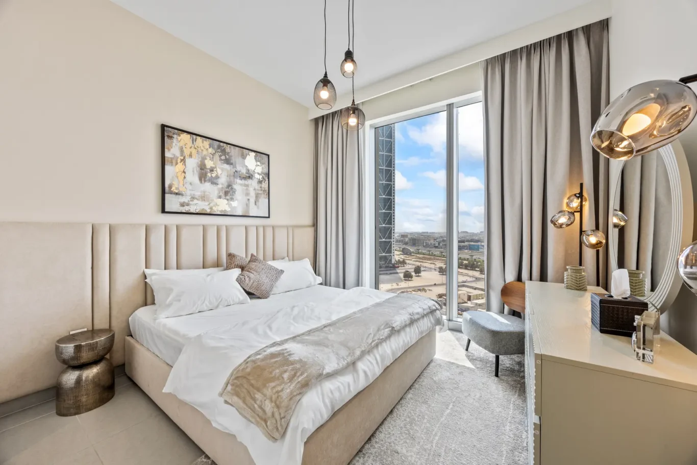A modern hotel bedroom with a padded headboard, white bedding, and beige throw pillows, beside a large window with gray curtains.