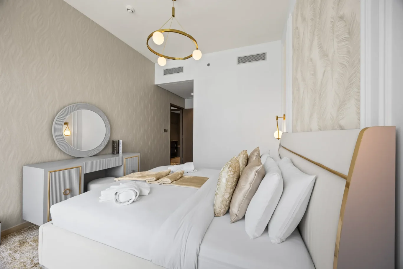 Two-bed hotel room with white linens, beige and gold pillows, and a circular gold chandelier above a white vanity with a round mirror on a textured wall.
