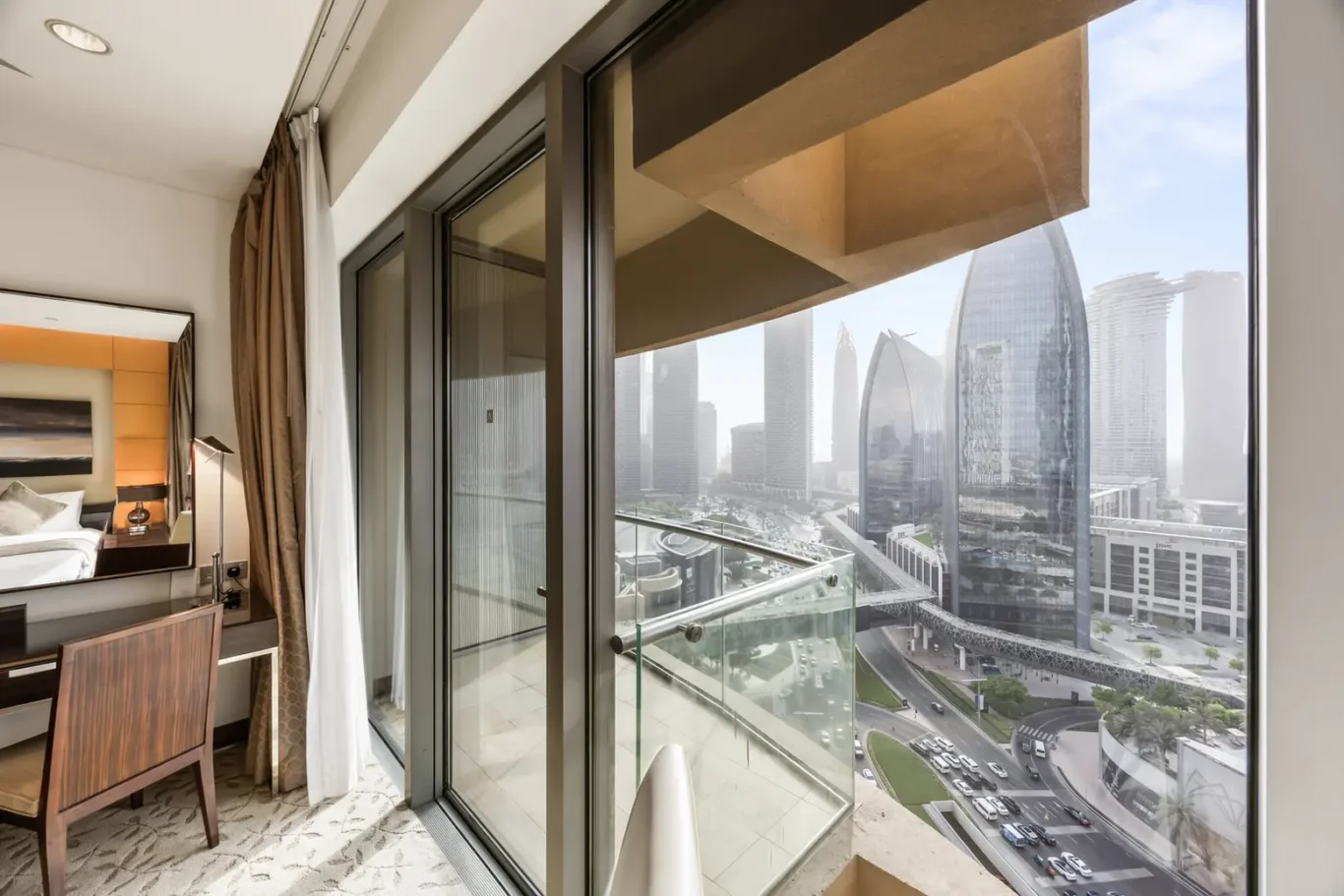 Hotel room with a glass balcony door, overlooking a futuristic cityscape with tall curved skyscrapers and a busy street below.