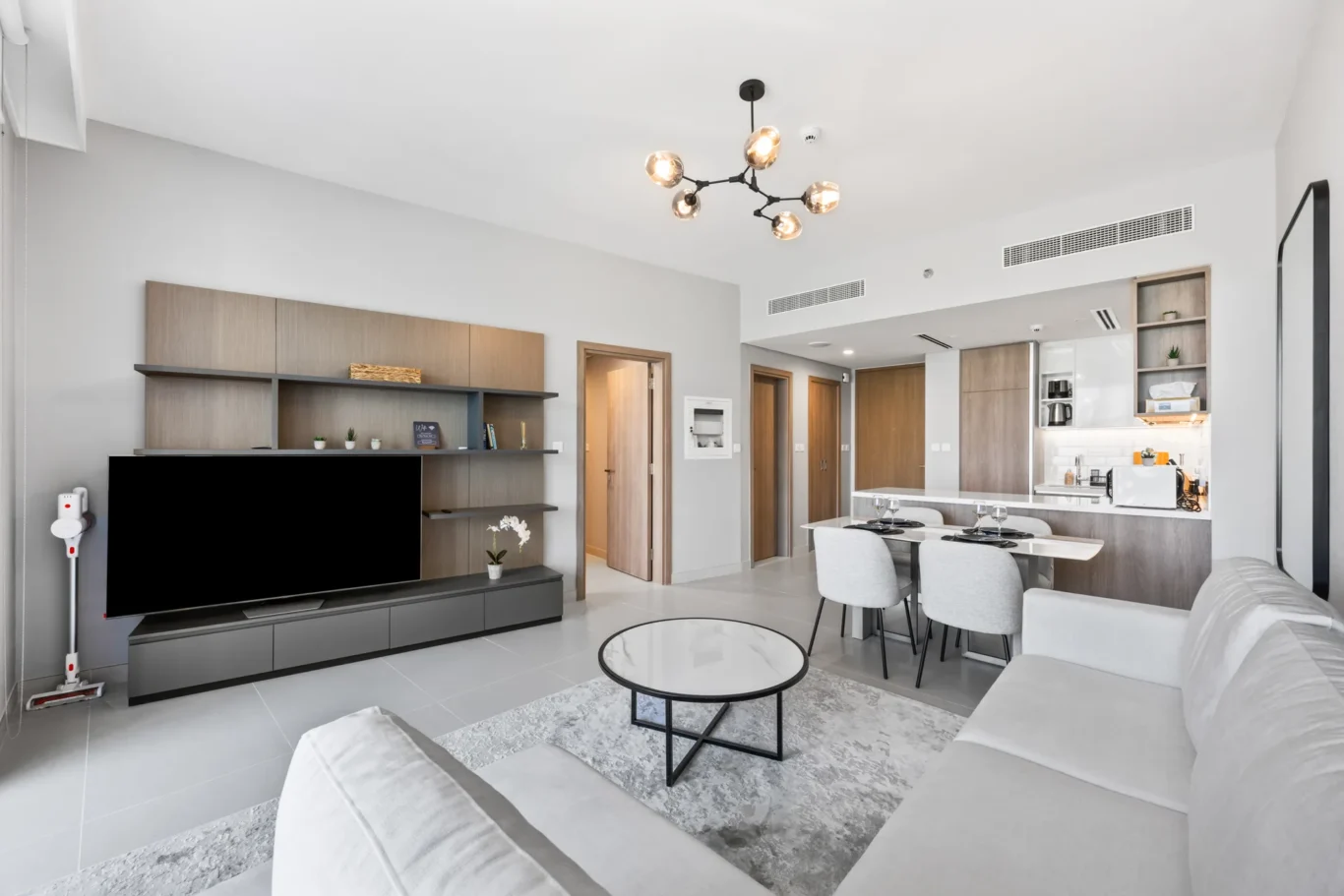 Open-plan living area with a wall-mounted TV, shelving unit, and a kitchen visible in the background.
