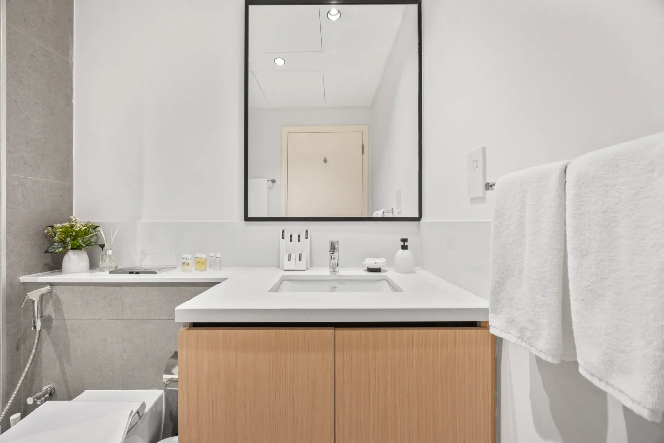 Modern bathroom vanity: white countertop with undermount sink, wooden cabinet, large framed mirror, and a white towel on the right edge.
