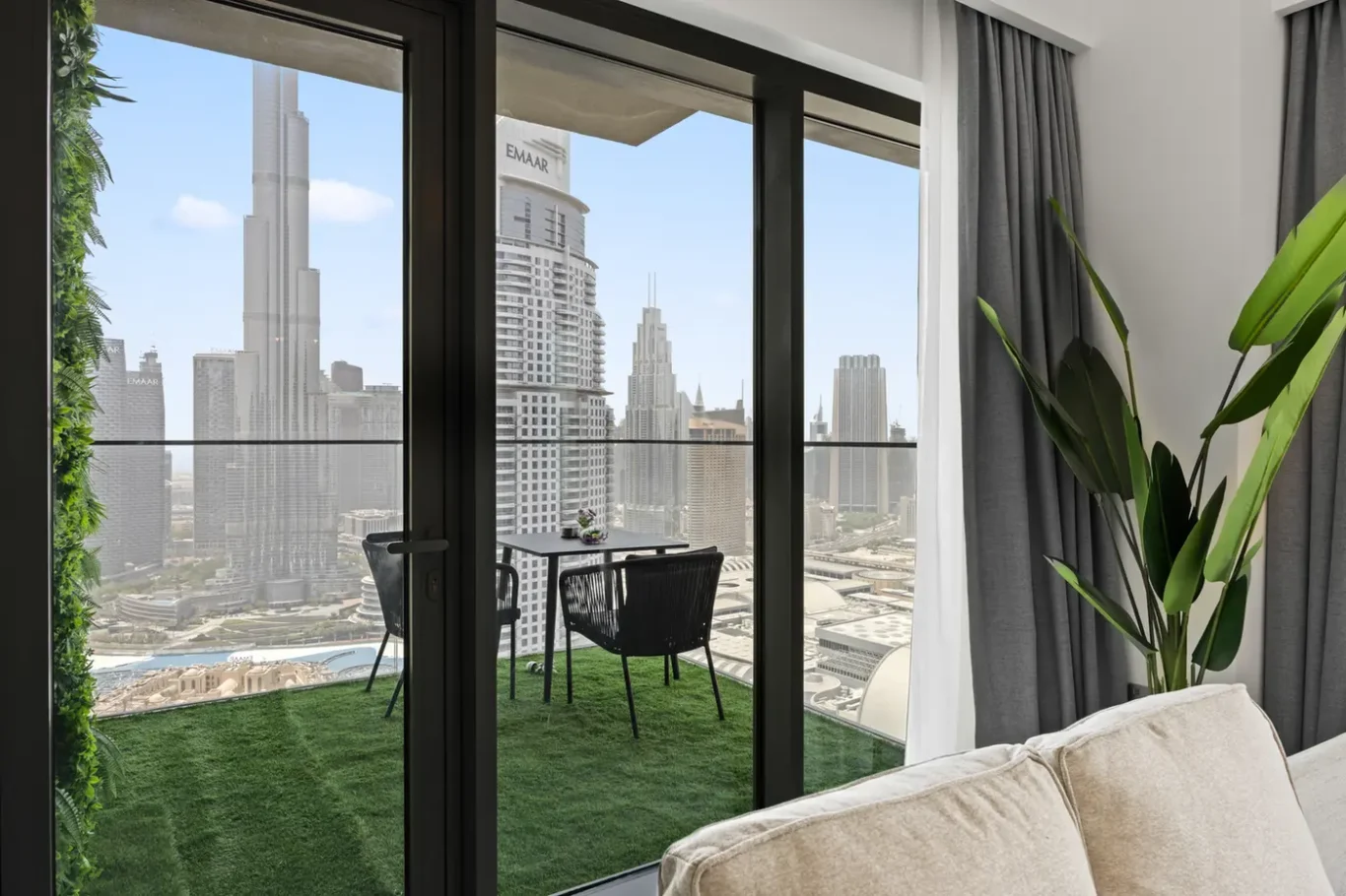Living room with glass doors opening to a small balcony, overlooking a city skyline of tall skyscrapers and a green artificial lawn outside.