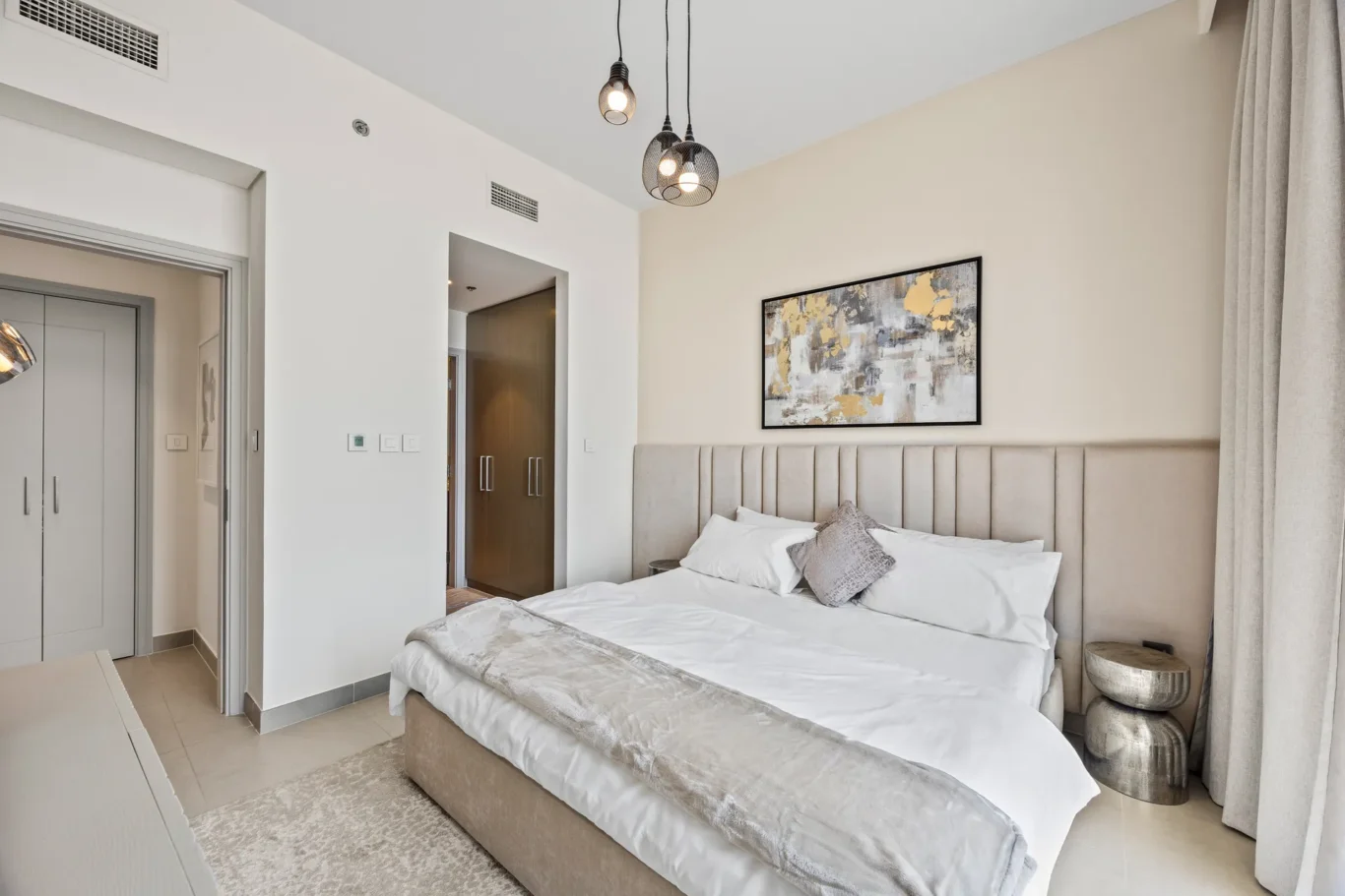 Bedroom with a padded beige headboard, white bedding, and abstract wall art; pendant lights hang from the ceiling above the bed.
