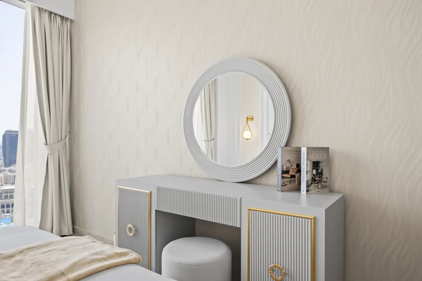 Grey vanity with ribbed doors and gold trim, topped by a large round silver-framed mirror, in a beige textured-wall bedroom with cream curtains and city view through a window on the left. Magazines sit on the dresser.
