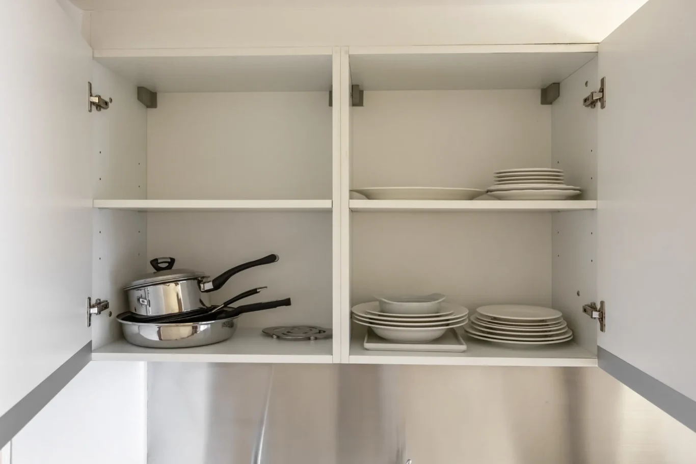 Open white kitchen cabinet with cookware on the lower left shelf and stacks of plates and bowls on the right shelves.