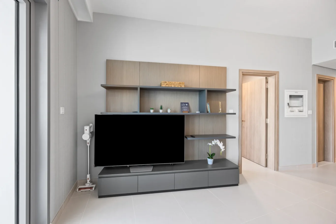 Modern living room with a gray entertainment unit and open shelves, a large flat-screen TV, and small potted plants.