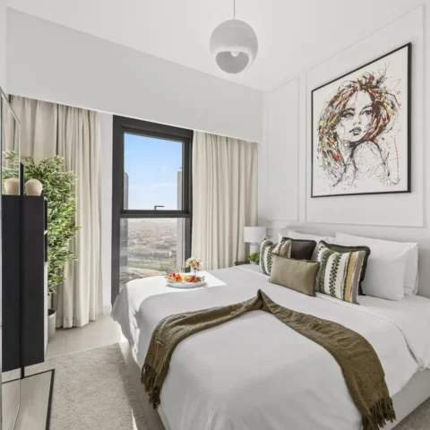 Modern hotel bedroom with large window, beige curtains, and a bed dressed in white linens with green-brown throw; abstract portrait hangs above. A tray of fruit sits on the bed.