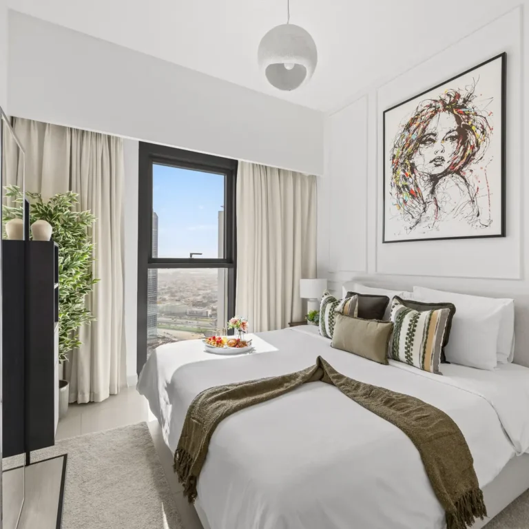 Modern hotel bedroom with large window, beige curtains, and a bed dressed in white linens with green-brown throw; abstract portrait hangs above. A tray of fruit sits on the bed.