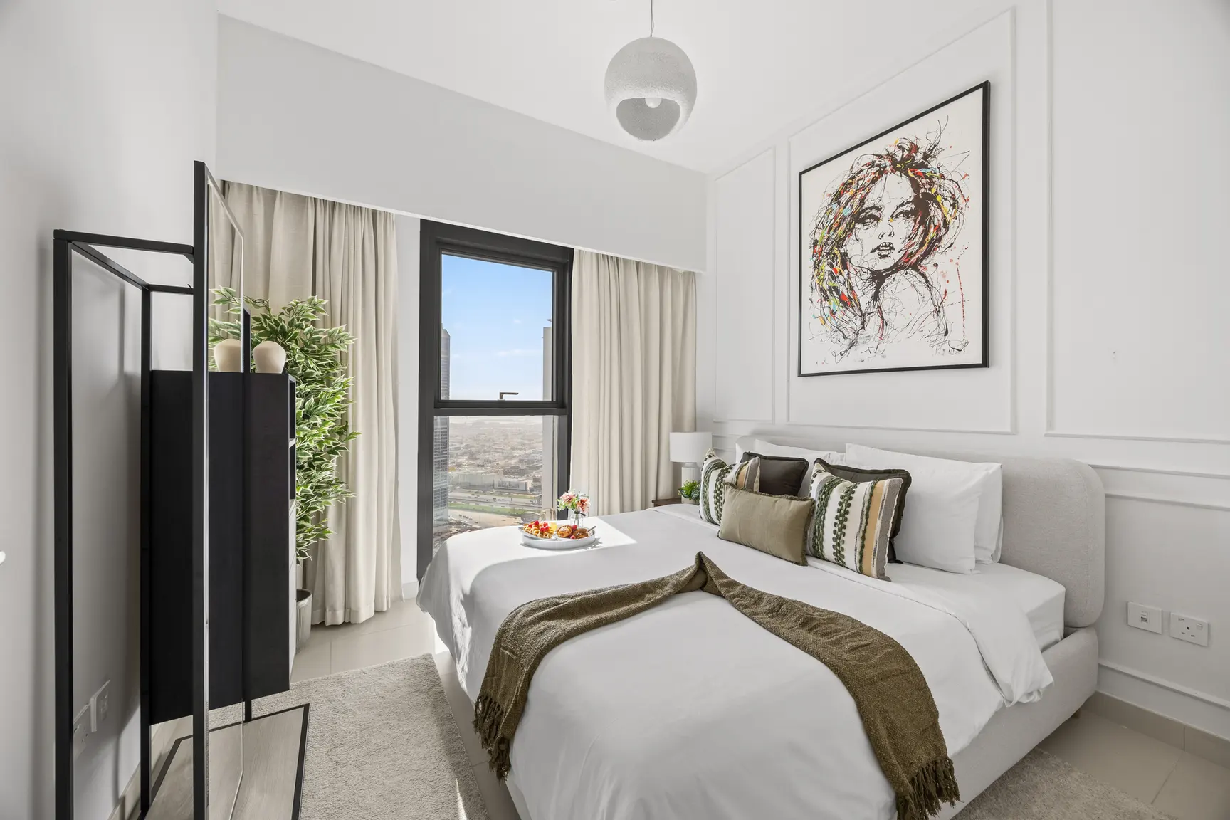 Modern hotel bedroom with large window, beige curtains, and a bed dressed in white linens with green-brown throw; abstract portrait hangs above. A tray of fruit sits on the bed.
