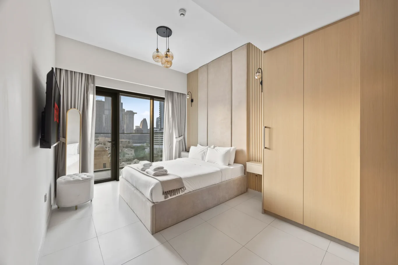 Modern hotel room with a king bed, beige upholstered headboard, and large glass doors to a city view. Neutral tones, pendant lights, and a tall wooden wardrobe.