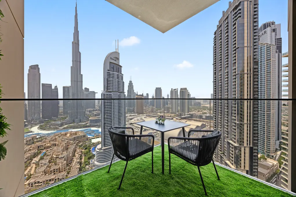 Balcony seating on artificial grass with a table and two chairs, overlooking a city skyline of tall skyscrapers under a clear blue sky.