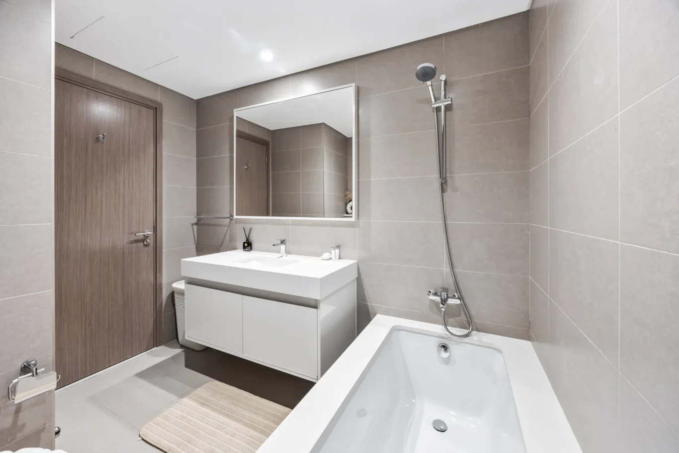 Modern bathroom with a white vanity, large rectangular mirror, and beige-tiled walls. A shower and bathtub are visible.