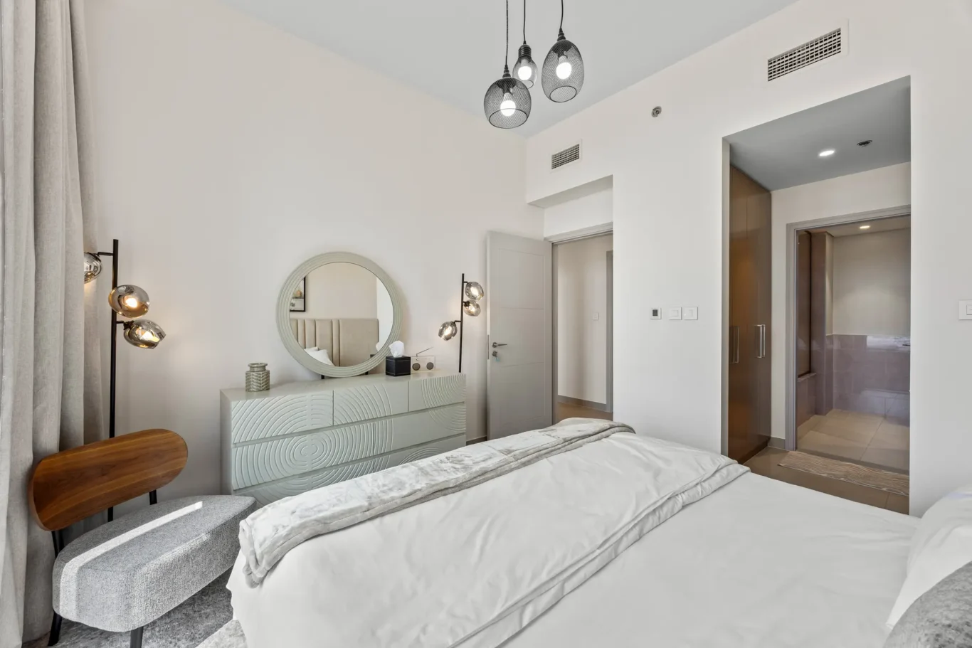 Modern bedroom with white decor: round mirror above a patterned dresser, bedside lamps, and grey curtains.