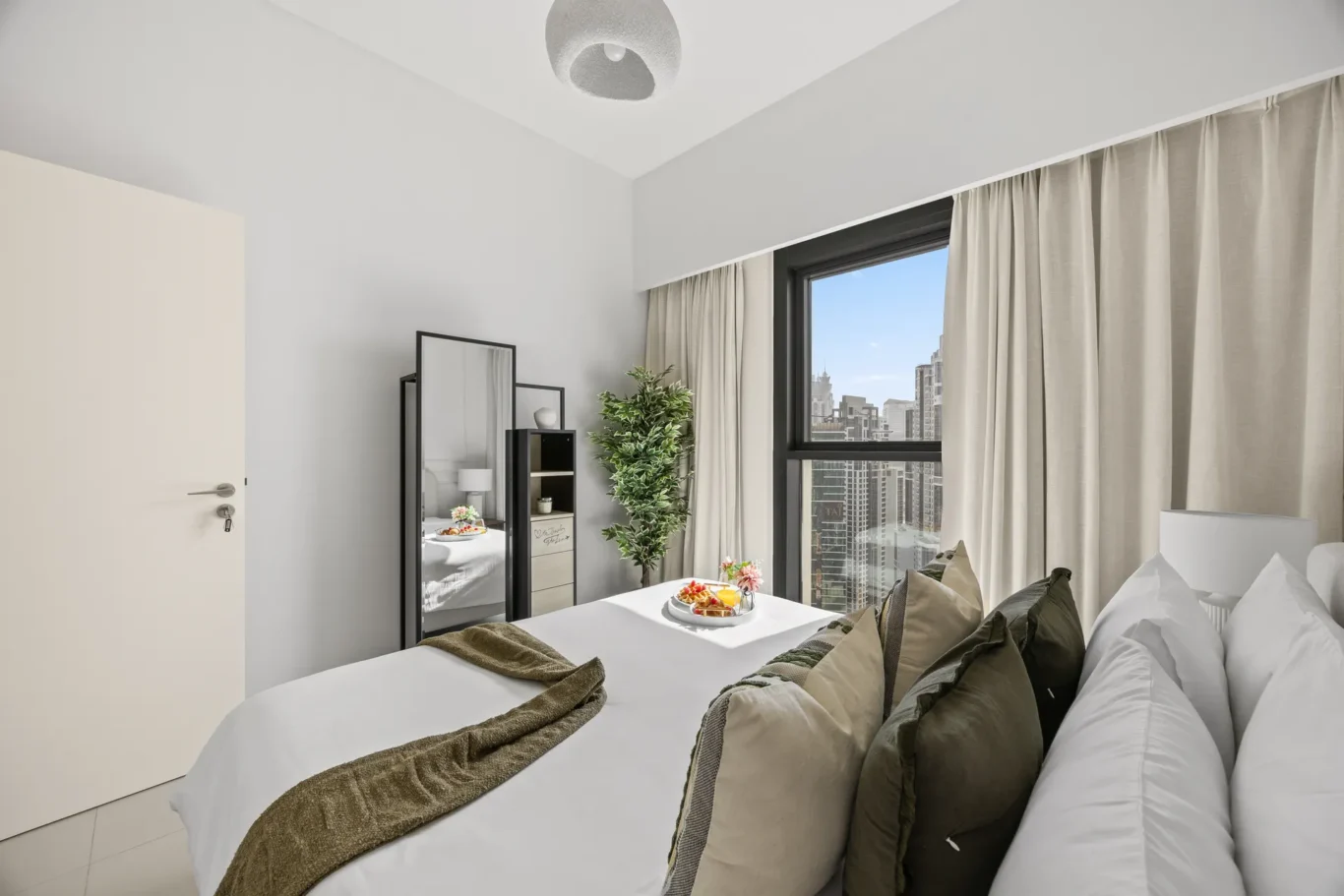 Bright hotel bedroom with a white bed, brown-and-taupe throw and pillows, and a city view through a large window with beige curtains. A breakfast tray and plant sit near the window.