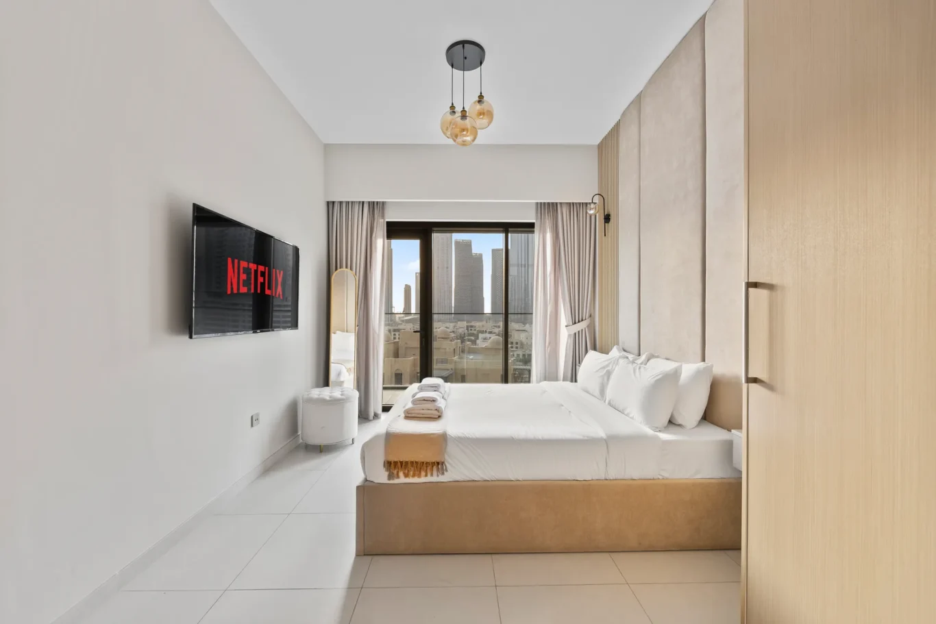 Bedroom in a modern hotel with a large white bed, folded towels, and a wall-mounted Netflix TV; balcony doors reveal a city skyline outside.