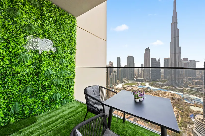 Balcony with a green vertical garden and neon 'Safe Stay Holiday Inn' sign, overlooking Dubai's skyline and Burj Khalifa.
