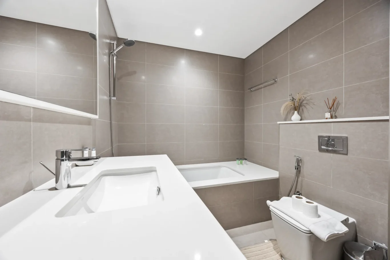 Contemporary grey-tiled bathroom with a white countertop sink, built-in bathtub, and chrome fixtures.