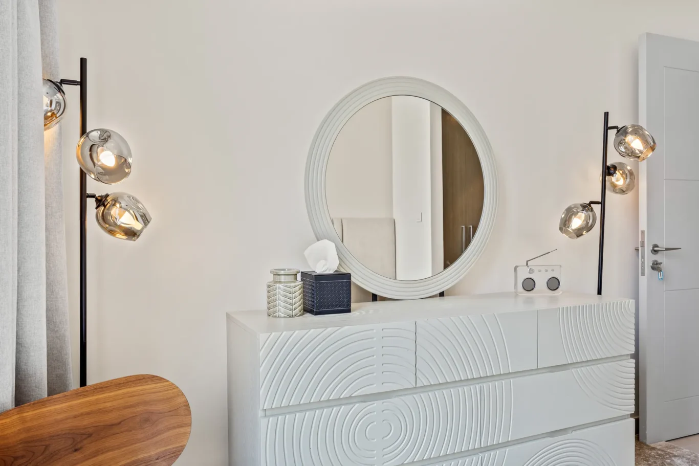 White dresser with a large circular mirror and glass globe lamps on either side, forming a modern vanity setup.
