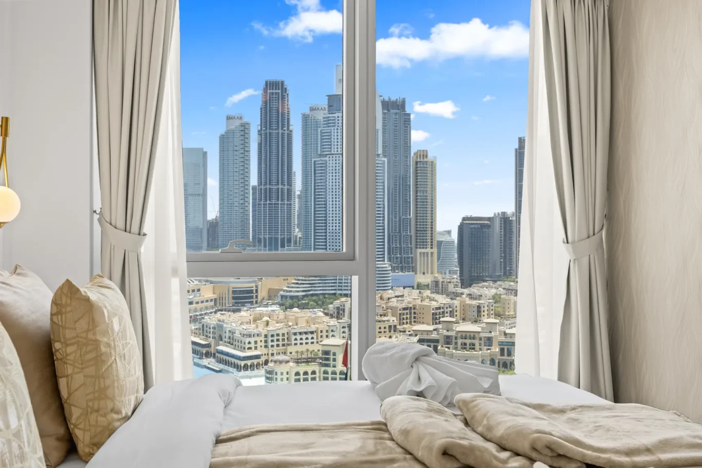 Hotel bedroom with a large window showing a city skyline; bed with beige pillows and cream blanket in foreground, curtains pulled back.