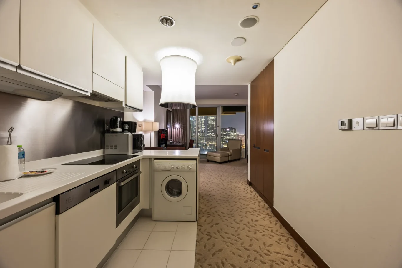 Bright, modern kitchen with white cabinets and stainless-steel appliances, plus a washer-dryer; city lights visible through the balcony doors in the background.